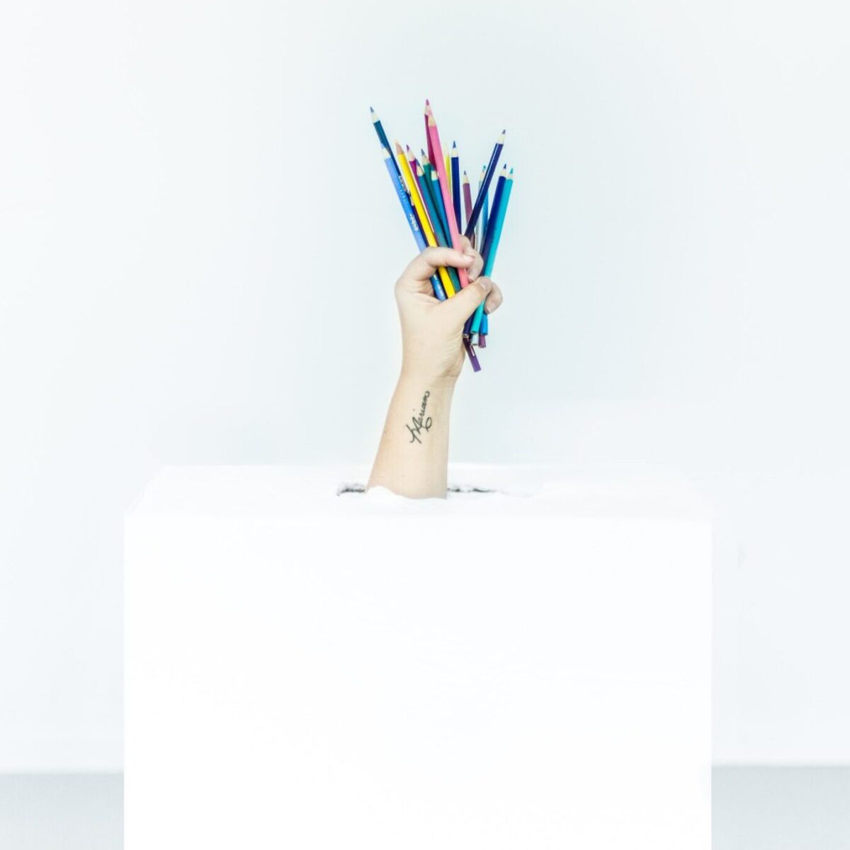 Tattooed hand emerging from a white box holding up different colored pencils