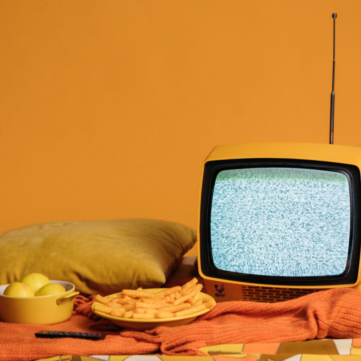 Small yellow TV showing static on a orange blanket next to a yellow pillow inside a room with yellow walls
