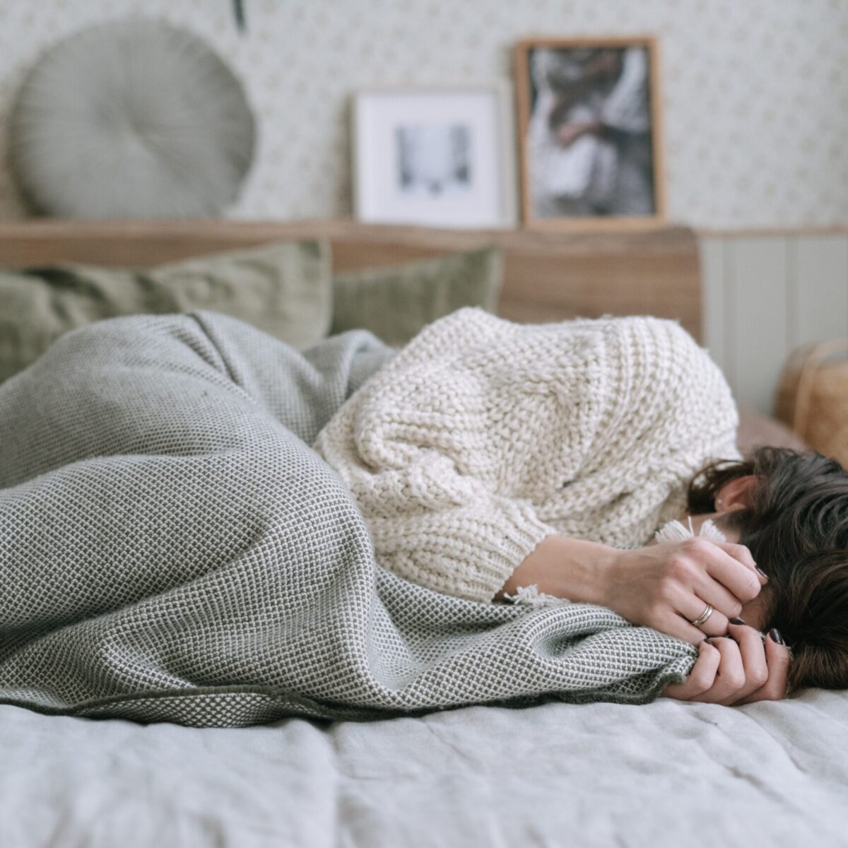 Brunette woman wearing sweatpants and a sweater laying on a bed in the fetal position